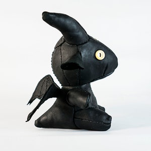 May include: Black leather plush toy shaped like a dragon with wings, horns, and a single yellow button eye.