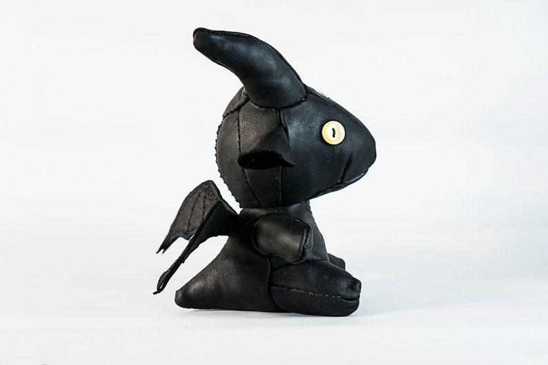 May include: Black leather plush toy shaped like a dragon with wings, horns, and a single yellow button eye.