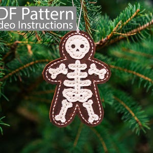 Könnte beinhalten: Ein brauner Filz-Skelett-Anhänger mit weißen Nähten hängt an einem grünen Kiefernbaumzweig. Der Anhänger ist in Form eines Lebkuchenmanns.  PDF Pattern Video Instructions.