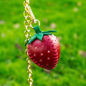 May include: A red leather strawberry keychain with green leaves and white stitching. The keychain is attached to a gold chain.