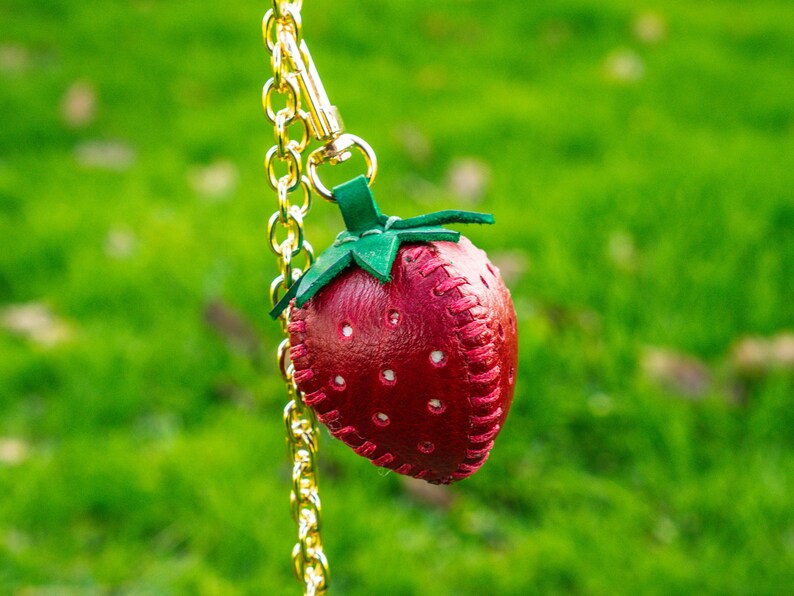 May include: A red leather strawberry keychain with green leaves and white stitching. The keychain is attached to a gold chain.