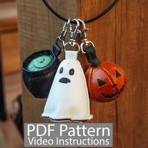 Puede incluir: Un conjunto de amuletos de cuero con temática de Halloween que presenta un caldero negro con contenido verde, un fantasma blanco y una calabaza naranja. Los amuletos están sujetos a un cordón negro con cierres plateados. El texto "PDF Pattern Video Instructions" es visible.