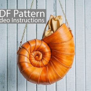 Op de afbeelding: Een bruine leren handtas in de vorm van een nautilus-schelp met een gouden kettingband. De tas hangt aan een witte houten muur.  PDF Pattern Video Instructions.