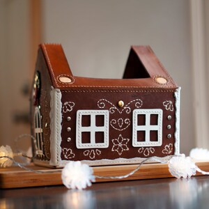 May include: A brown leather house-shaped box with white stitched details, resembling a gingerbread house. Features two white window cutouts and decorative stitching. A string of white lights is draped in front of the house. The house is on a wooden surface.