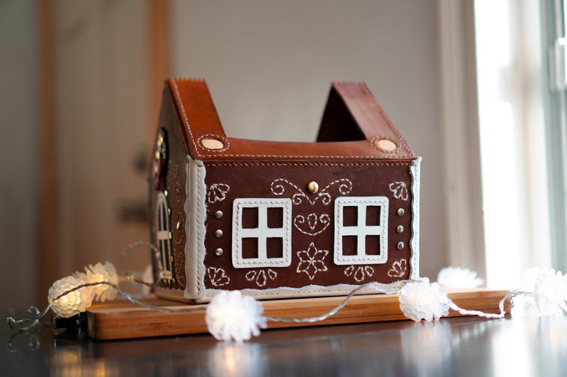May include: A brown leather house-shaped box with white stitched details, resembling a gingerbread house. Features two white window cutouts and decorative stitching. A string of white lights is draped in front of the house. The house is on a wooden surface.