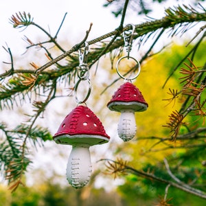 Peut inclure: Deux porte-cl&eacute;s en cuir en forme de champignon rouge et blanc suspendus &agrave; une branche d'arbre. Les porte-cl&eacute;s ont une tige blanche et un chapeau rouge avec des coutures blanches.