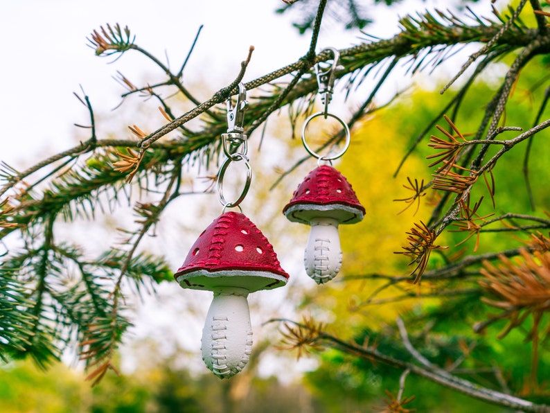 Peut inclure: Deux porte-cl&eacute;s en cuir en forme de champignon rouge et blanc suspendus &agrave; une branche d'arbre. Les porte-cl&eacute;s ont une tige blanche et un chapeau rouge avec des coutures blanches.