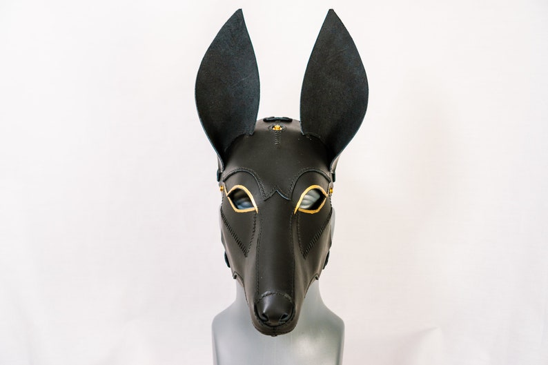 May include: A black leather mask with gold accents, featuring large ears and a pointed snout. The mask is designed to cover the entire face, with eye holes and a small opening for the mouth.