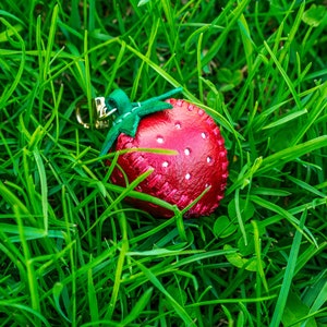 May include: A red leather strawberry keychain with a green leather stem and leaves. The keychain is lying on a bed of green grass.