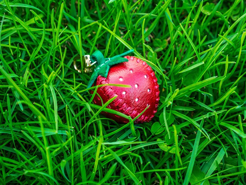 May include: A red leather strawberry keychain with a green leather stem and leaves. The keychain is lying on a bed of green grass.