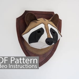 May include: A raccoon head wall mount made of fabric, with a brown leather-like shield. The raccoon has a white face, black eyes, and a black nose. The ears are brown. The text "PDF Pattern Video Instructions" is at the bottom.