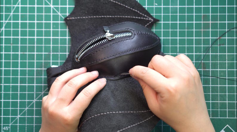 May include: A close-up of a leather craft project in progress. A black leather pouch with a silver zipper is being sewn onto a dark gray fabric, possibly felt, with a needle and thread. The background is a green cutting mat.