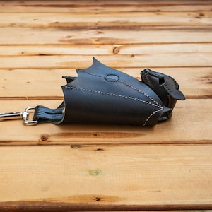 May include: Black leather dragon-shaped pouch with white stitching and a silver-colored clasp. The pouch has a wing-like design and a small head detail. The leather is a matte finish, and the pouch is lying on a wooden surface.