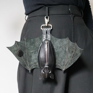 May include: A black leather bat-shaped bag with a zipper closure and silver-tone hardware. The bag has bat wing details and is attached to a silver clip. The bag is hanging from a black belt.