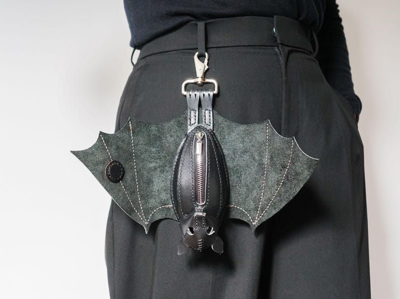 May include: A black leather bat-shaped bag with a zipper closure and silver-tone hardware. The bag has bat wing details and is attached to a silver clip. The bag is hanging from a black belt.