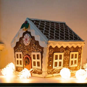 May include: A decorative gingerbread house with brown walls, a white roof, and white trim. The house features white embroidered details, a red door, and a candy cane design. A string of white lights is in front of the house.