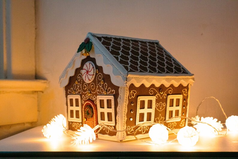 May include: A decorative gingerbread house with brown walls, a white roof, and white trim. The house features white embroidered details, a red door, and a candy cane design. A string of white lights is in front of the house.