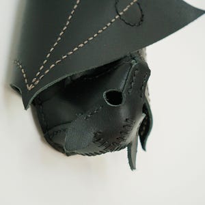 May include: A black leather bat-shaped mask with tan stitching. The mask has pointed ears and a stitched nose and mouth. The wings are folded over the top of the mask. The mask is on a white background.