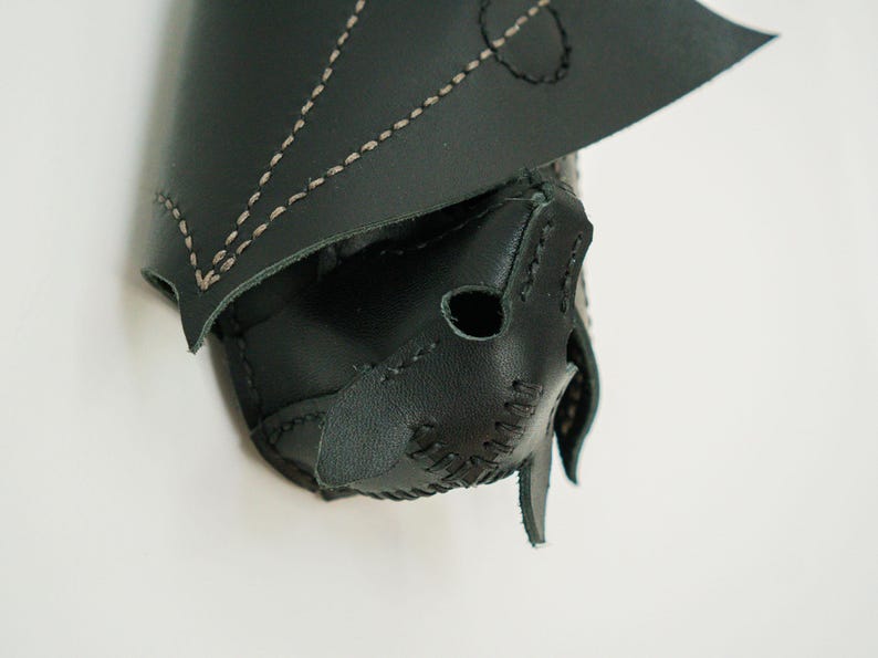 May include: A black leather bat-shaped mask with tan stitching. The mask has pointed ears and a stitched nose and mouth. The wings are folded over the top of the mask. The mask is on a white background.