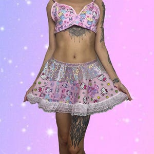 May include: A pink two-piece lingerie set featuring a bra and a skirt. The bra has thin straps and a bow. The skirt has a sparkly top layer and a pink layer with a cartoon character print, trimmed with white lace.