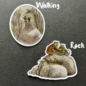 Puede incluir: Dos pegatinas de ranas, una con dos ranas caminando por un sendero a trav&eacute;s de hierba alta y la otra con dos ranas sentadas en una gran roca.