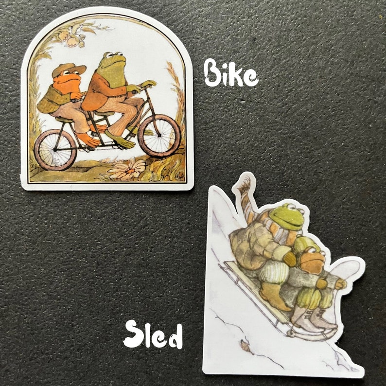 Puede incluir: Dos ranas montando una bicicleta t&aacute;ndem con un cuadro blanco y un manillar marr&oacute;n. Las ranas llevan chaquetas marrones y la bicicleta est&aacute; en un camino con hierba verde y flores.