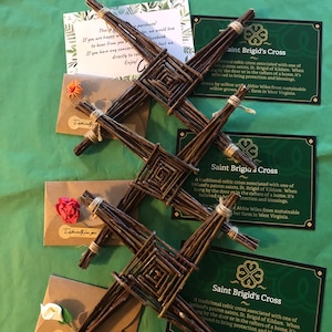Op de afbeelding: Drie Saint Brigid's kruisen gemaakt van geweven wilgentakken, weergegeven met cadeaukaarten en kleine enveloppen. De kruisen zijn bruin, met een vierkant midden en vier uitstralende armen. De cadeaukaarten zijn groen met witte tekst en een klaverlogo.