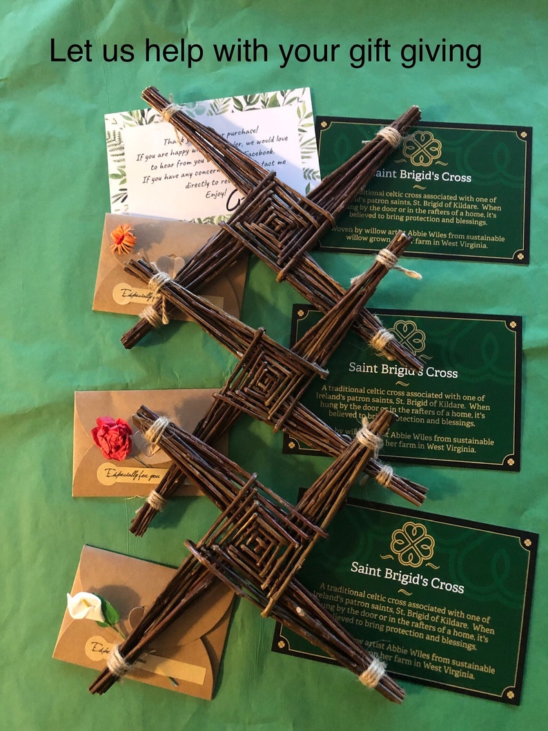 Op de afbeelding: Drie Saint Brigid's kruisen gemaakt van geweven wilgentakken, weergegeven met cadeaukaarten en kleine enveloppen. De kruisen zijn bruin, met een vierkant midden en vier uitstralende armen. De cadeaukaarten zijn groen met witte tekst en een klaverlogo.