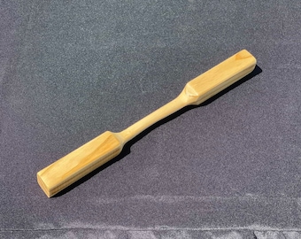 May include: A light-colored wooden stirring paddle featuring two rectangular ends connected by a slender, rounded central handle. The wood grain is visible throughout the smooth, polished surface of this kitchen utensil.