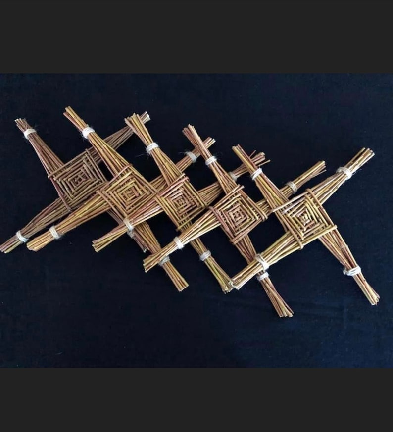 Op de afbeelding: Een verzameling geweven stro St. Brigid's kruisen, elk met een vierkant midden en vier uitstralende armen. De kruisen zijn vastgebonden met wit touw en diagonaal geplaatst tegen een zwarte achtergrond.