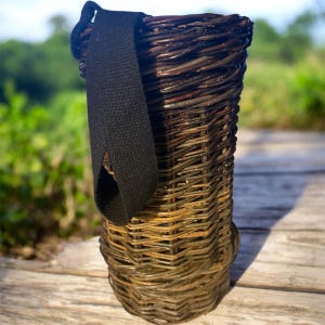 May include: A tall, cylindrical basket woven from dark brown willow branches, featuring a sturdy black fabric strap attached to the top rim. The woven structure displays a tight, vertical pattern with visible natural variations in the wood grain.