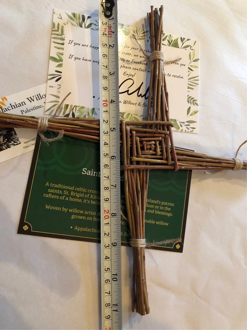 Op de afbeelding: Een handgemaakt Sint-Brigidakruis van gevlochten wilgentakken, ongeveer 25 cm lang. Het kruis heeft een vierkant middenontwerp en is vastgebonden met touw. Inclusief een label met tekst.