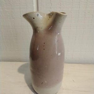 以下が含まれることがあります： ユニークな非対称の縁を持つ手作りのセラミック花瓶。花瓶は、明るいベージュのトップと柔らかく落ち着いたモーブのボディを備えた2色の配色です。ベースはクリーミーホワイトです。花瓶の高さは約18cmです。