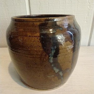 Puede incluir: Un jarrón de cerámica marrón con un esmalte marrón oscuro y una raya azul claro que recorre el lateral.
