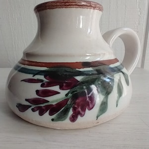 Puede incluir: Una taza de cer&aacute;mica blanca con un borde marr&oacute;n y un dise&ntilde;o floral. Las flores est&aacute;n pintadas en tonos de morado, verde y marr&oacute;n.