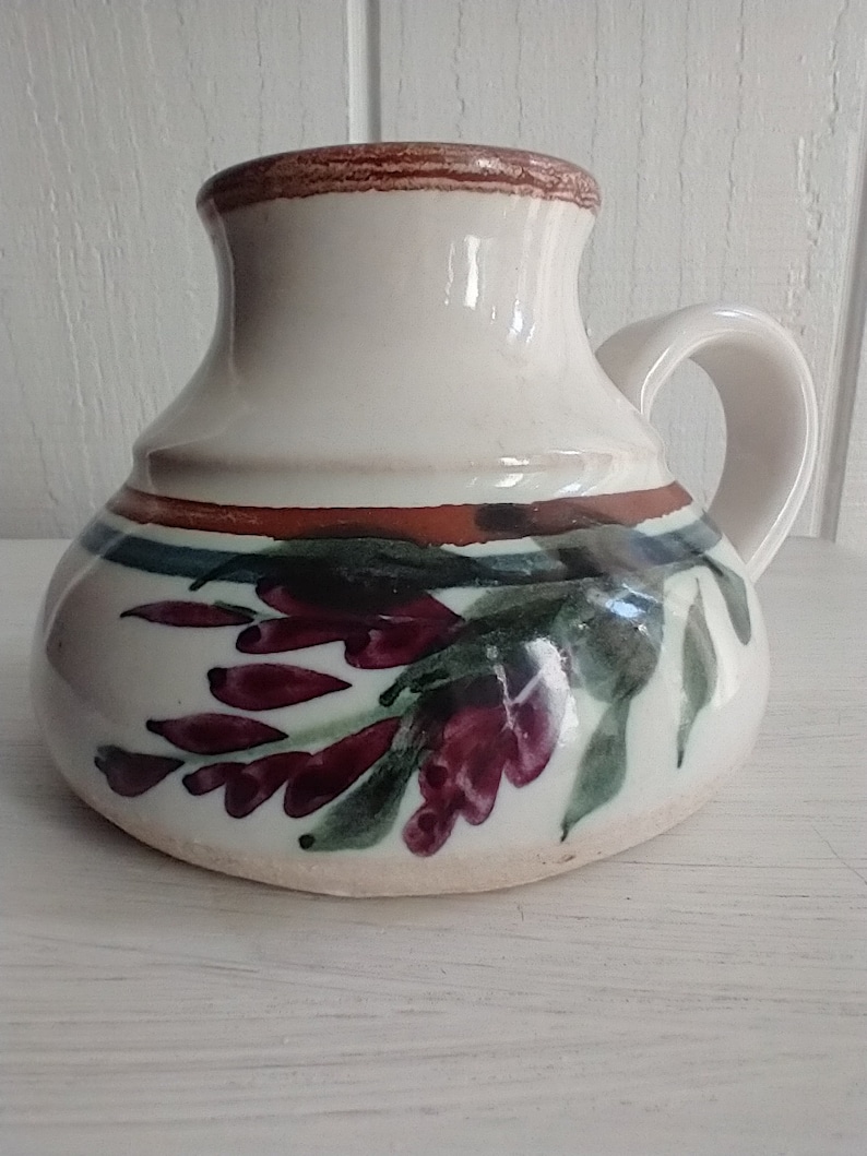 Puede incluir: Una taza de cer&aacute;mica blanca con un borde marr&oacute;n y un dise&ntilde;o floral. Las flores est&aacute;n pintadas en tonos de morado, verde y marr&oacute;n.