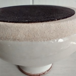 Puede incluir: Taz&oacute;n de pedestal de cer&aacute;mica blanca con una base de fieltro marr&oacute;n. El taz&oacute;n tiene un acabado liso y brillante y una forma ligeramente redondeada.