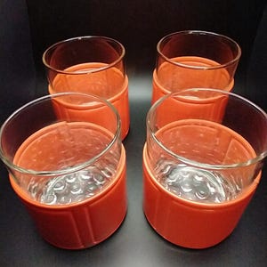 Op de afbeelding: Vier heldere drinkglazen met oranje plastic houders. De glazen hebben een textuurpatroon op de bodem. De oranje houders hebben een cilindrische vorm en zijn ontworpen om perfect in de glazen te passen.