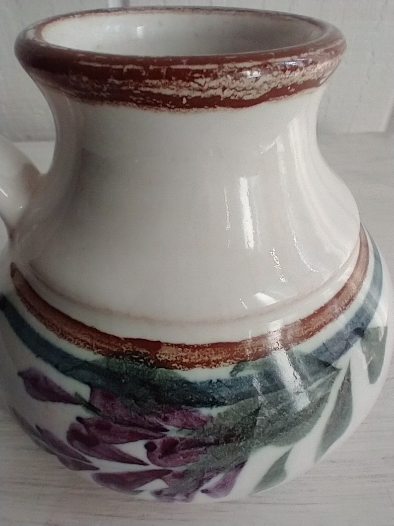 Puede incluir: Jarra de cer&aacute;mica blanca con un borde marr&oacute;n y un dise&ntilde;o floral. El dise&ntilde;o presenta flores moradas, hojas verdes y detalles marrones.