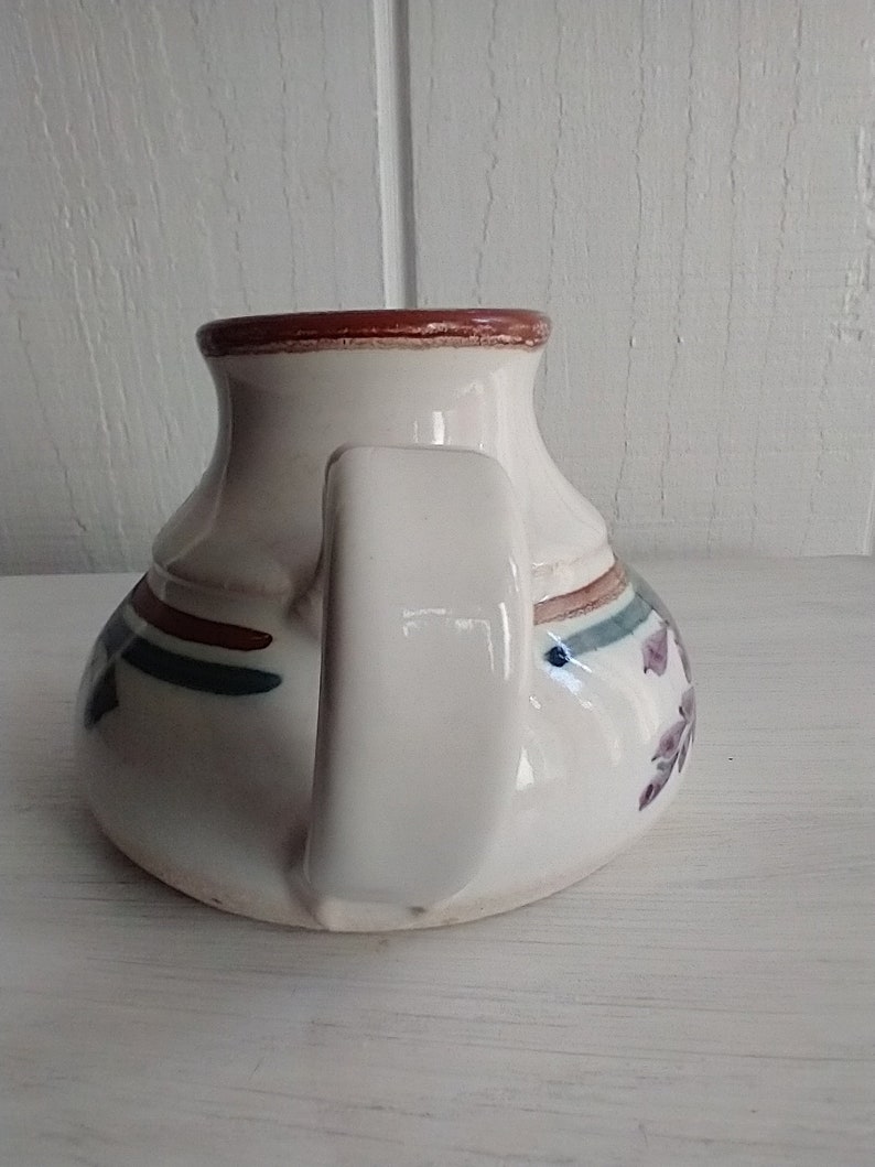 Puede incluir: Jarra de cer&aacute;mica blanca con un borde marr&oacute;n y un asa. La jarra tiene un dise&ntilde;o floral azul y verde con acentos rojos.