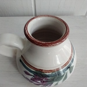 Puede incluir: Una jarra de cer&aacute;mica blanca con un borde marr&oacute;n y un dise&ntilde;o floral. La jarra tiene un asa y est&aacute; decorada con detalles en morado, verde y marr&oacute;n.