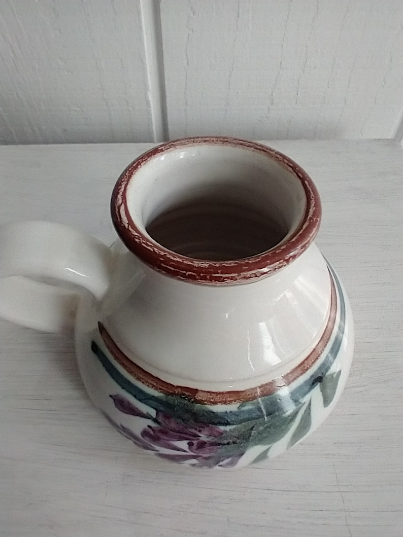 Puede incluir: Una jarra de cer&aacute;mica blanca con un borde marr&oacute;n y un dise&ntilde;o floral. La jarra tiene un asa y est&aacute; decorada con detalles en morado, verde y marr&oacute;n.