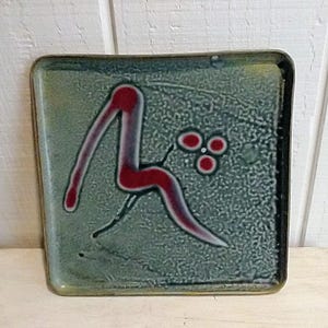 May include: Square ceramic tile with a green background and a red and white abstract design. The design features a curved line and three red circles.