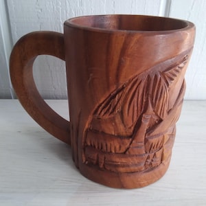 May include: A wooden mug with a carved design of a tropical scene. The mug is brown and has a handle.