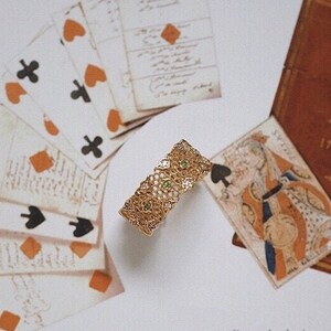 Può includere: Un braccialetto d'oro con un delicato design a filigrana e pietre preziose verdi. Il braccialetto è appoggiato su una superficie bianca con carte da gioco e un libro.