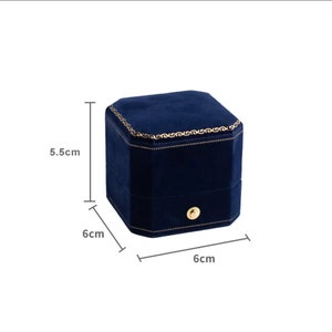 Op de afbeelding: Een blauwe fluwelen ringdoos met een gouden rand en een kleine knop aan de voorkant. De doos meet 6 cm bij 6 cm bij 5,5 cm.