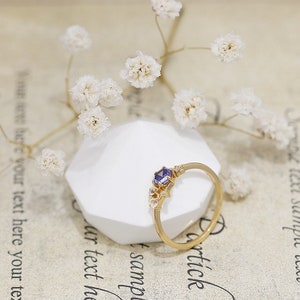 May include: A gold ring featuring a purple gemstone and small white gemstones. The ring is displayed on a white stand.