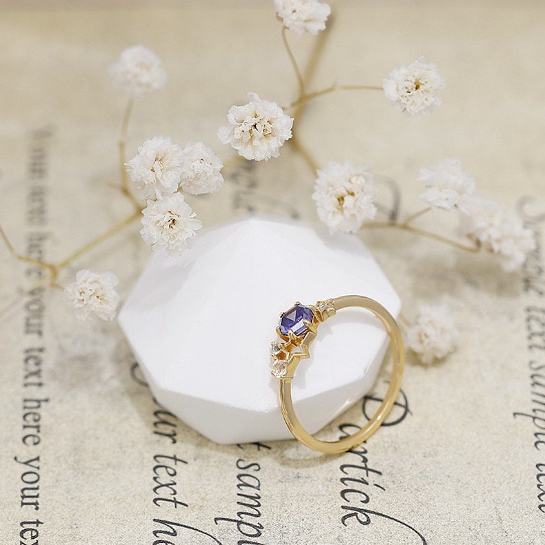 May include: A gold ring featuring a purple gemstone and small white gemstones. The ring is displayed on a white stand.