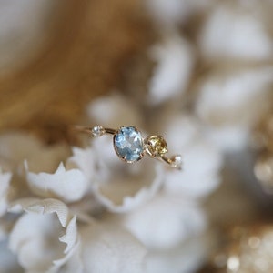 Könnte beinhalten: Ein zarter Goldring mit einem hellblauen Edelstein und einem kleineren gelben Edelstein. Der Ring hat ein geschwungenes Design mit kleinen Diamanten, die entlang der Ringschiene gesetzt sind.
