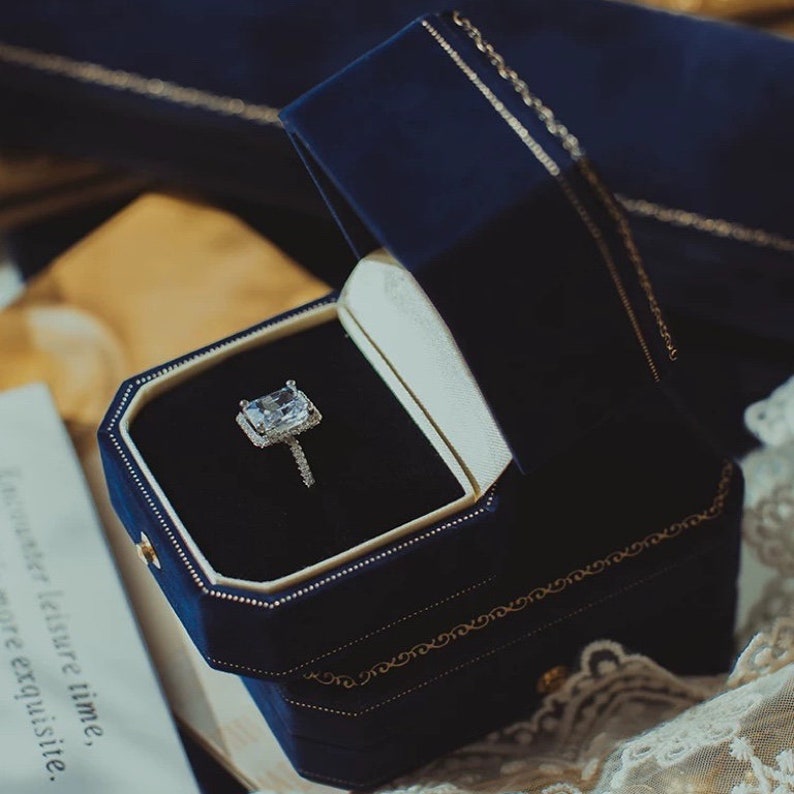 Op de afbeelding: Een vierkante ringdoos met een blauwe fluwelen binnenkant en een gouden rand. De doos is open en er ligt een diamanten ring in.